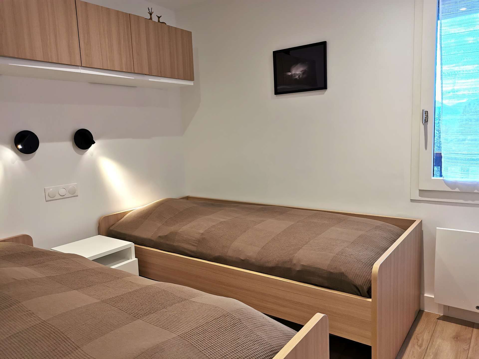 Une chambre avec deux lits, une armoire en bois, un tableau accroché au mur et une fenêtre avec des stores.