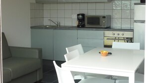 Eine kleine Küche mit grauen Schränken, einer Mikrowelle, einer Kaffeemaschine und einem Herd. Ein weißer Esstisch mit Stühlen und eine Couch stehen in der Ecke.