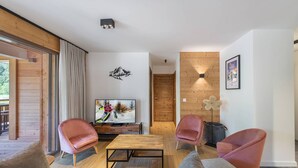 A modern living room with a wooden floor, a TV on a wooden stand, two pink chairs, a sofa, and a wooden wall with a decorative light fixture.