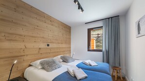 A modern bedroom with two beds, a wooden headboard, a window with curtains, and a small stool.