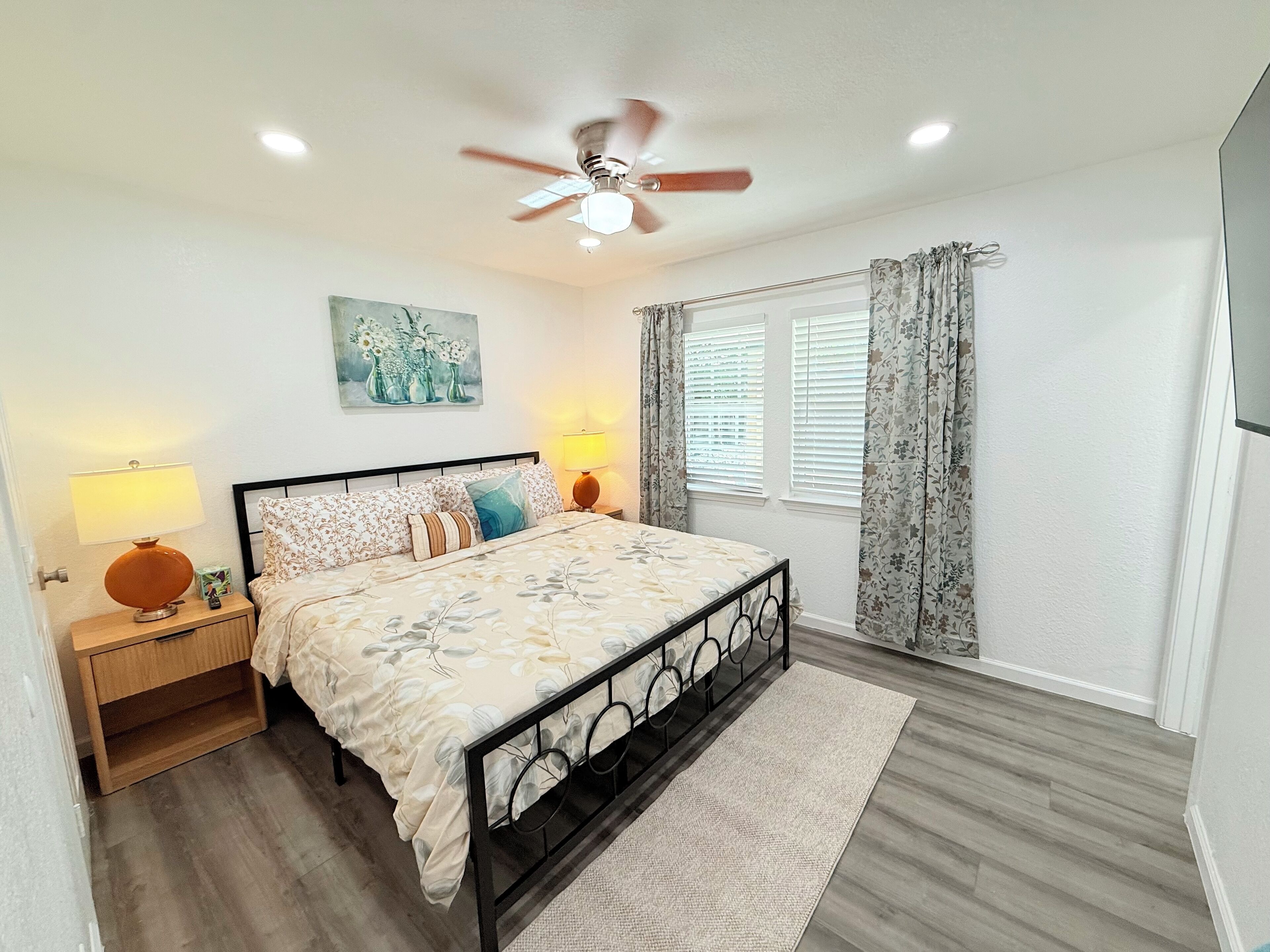 A bedroom with a bed, nightstands, a ceiling fan, a painting, and a window with curtains.