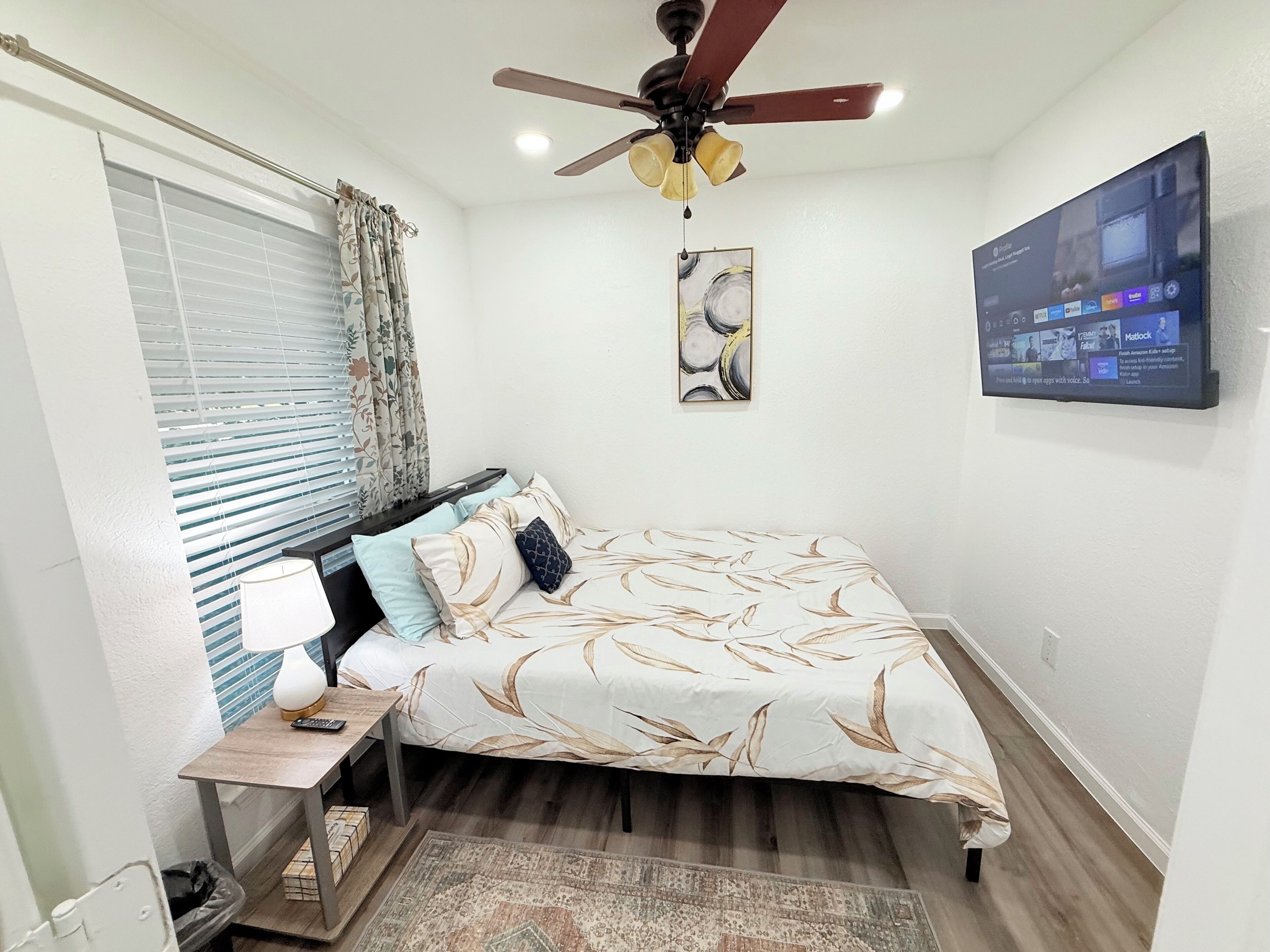 Une chambre à coucher comprenant un lit, une table de chevet, une lampe, un ventilateur de plafond, une fenêtre avec des stores, un tableau et un téléviseur fixé au mur.