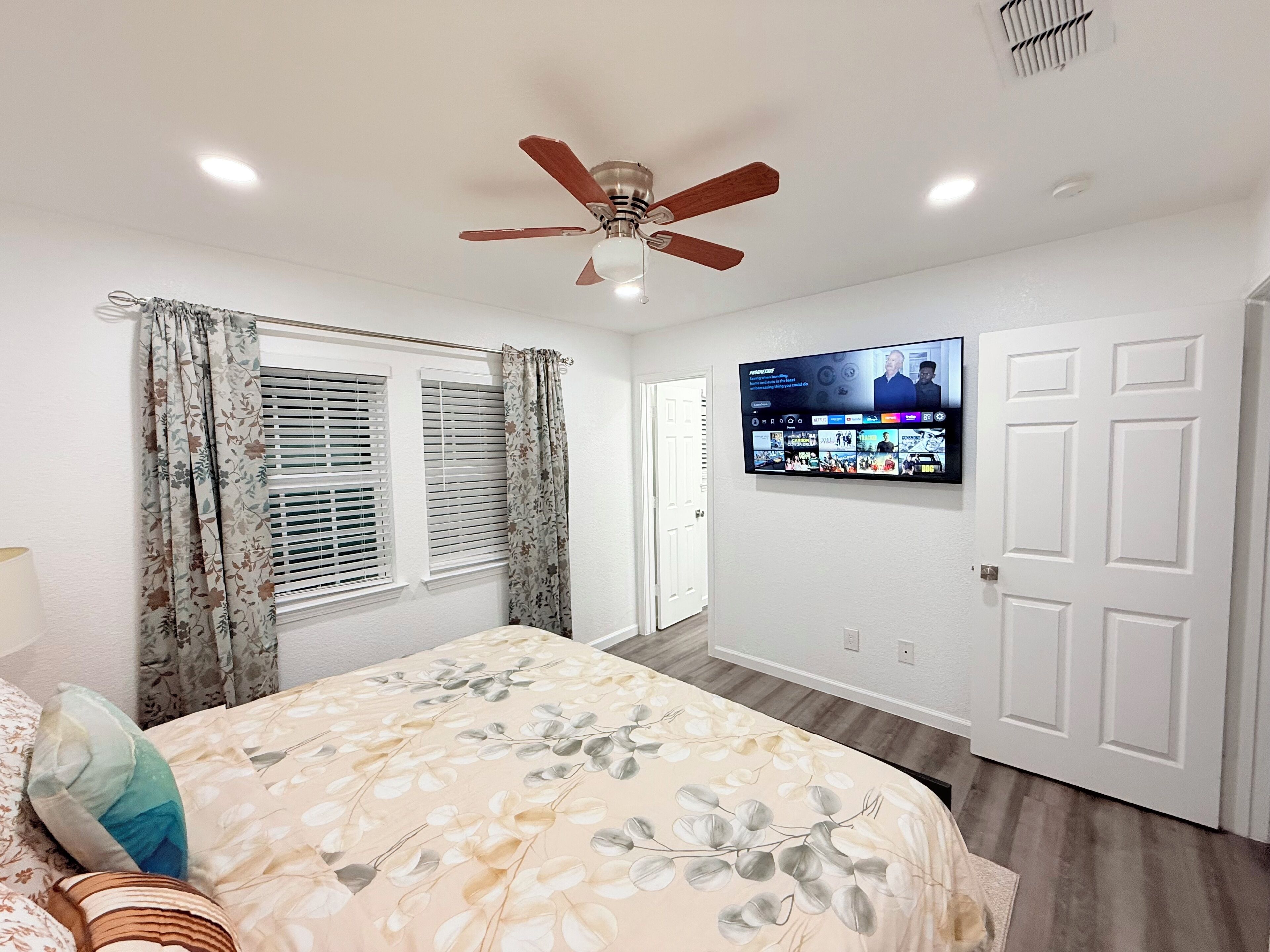 A bedroom with a bed, a ceiling fan, a mounted TV, and a door.