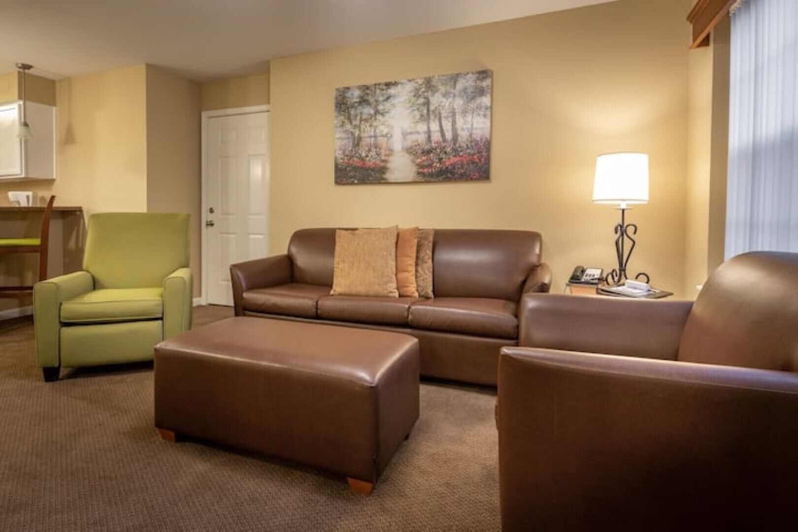 A hotel room with a brown leather sofa, a green armchair, and a brown ottoman. There is a painting on the wall and a lamp on a side table.