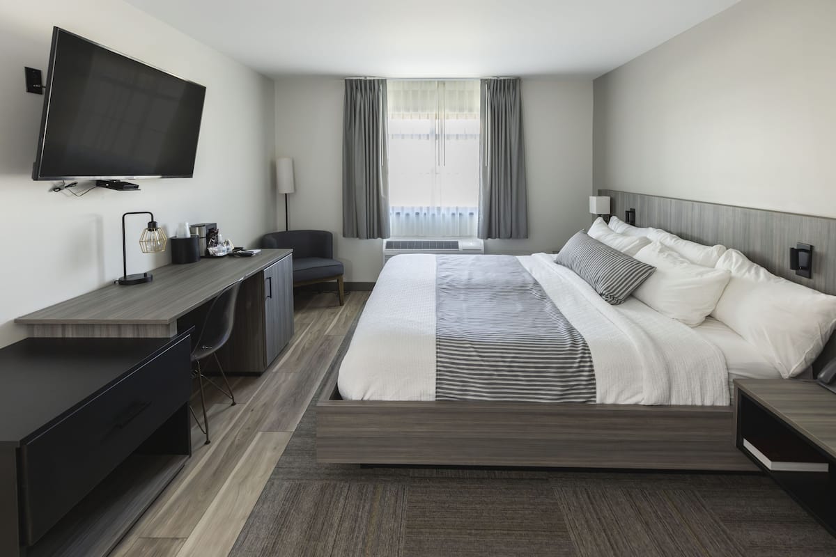 A modern hotel room with a flat-screen TV, a desk with a chair, a bed with multiple pillows, and a large window with curtains.