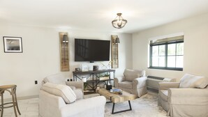 Una sala de estar con un televisor de pantalla plana grande, dos sofás color beige, una mesa de centro y una mesita de noche de madera.