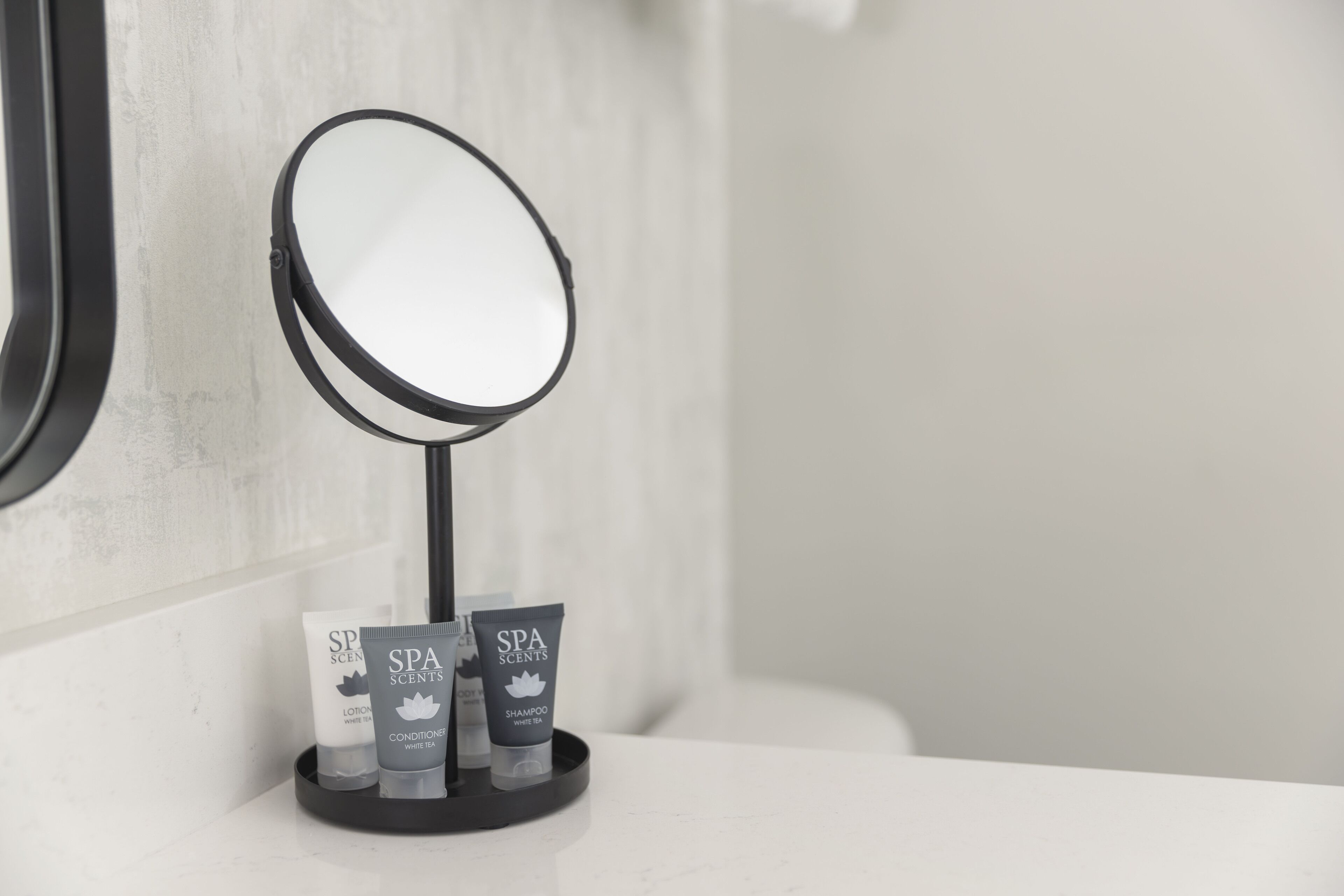Un meuble-lavabo de salle de bain avec un miroir rond, un plateau noir contenant trois produits de spa et un plan de toilette blanc.