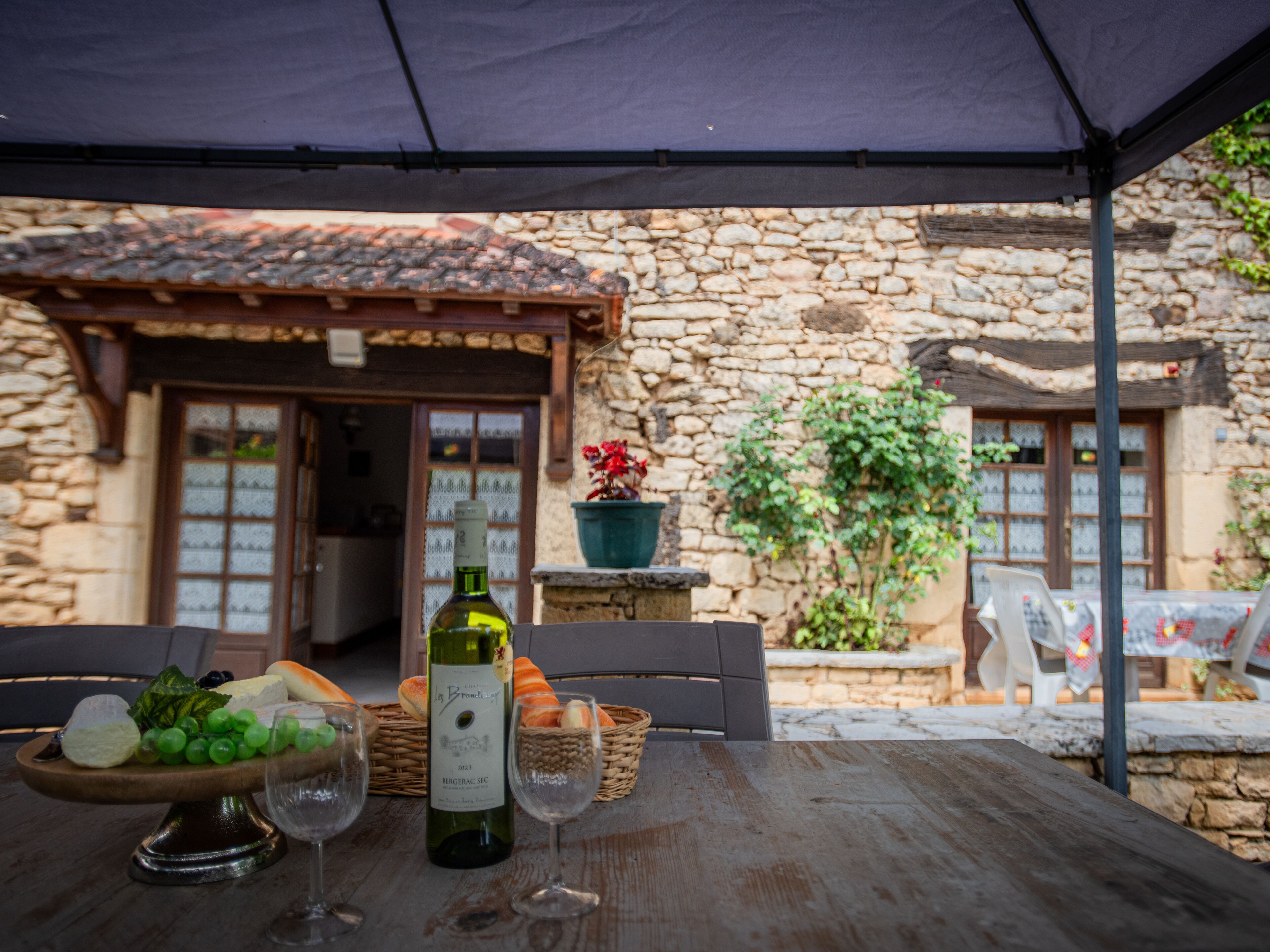 A rustic outdoor dining area with a stone building in the background, a table with a bottle of wine, glasses, and a basket of bread.