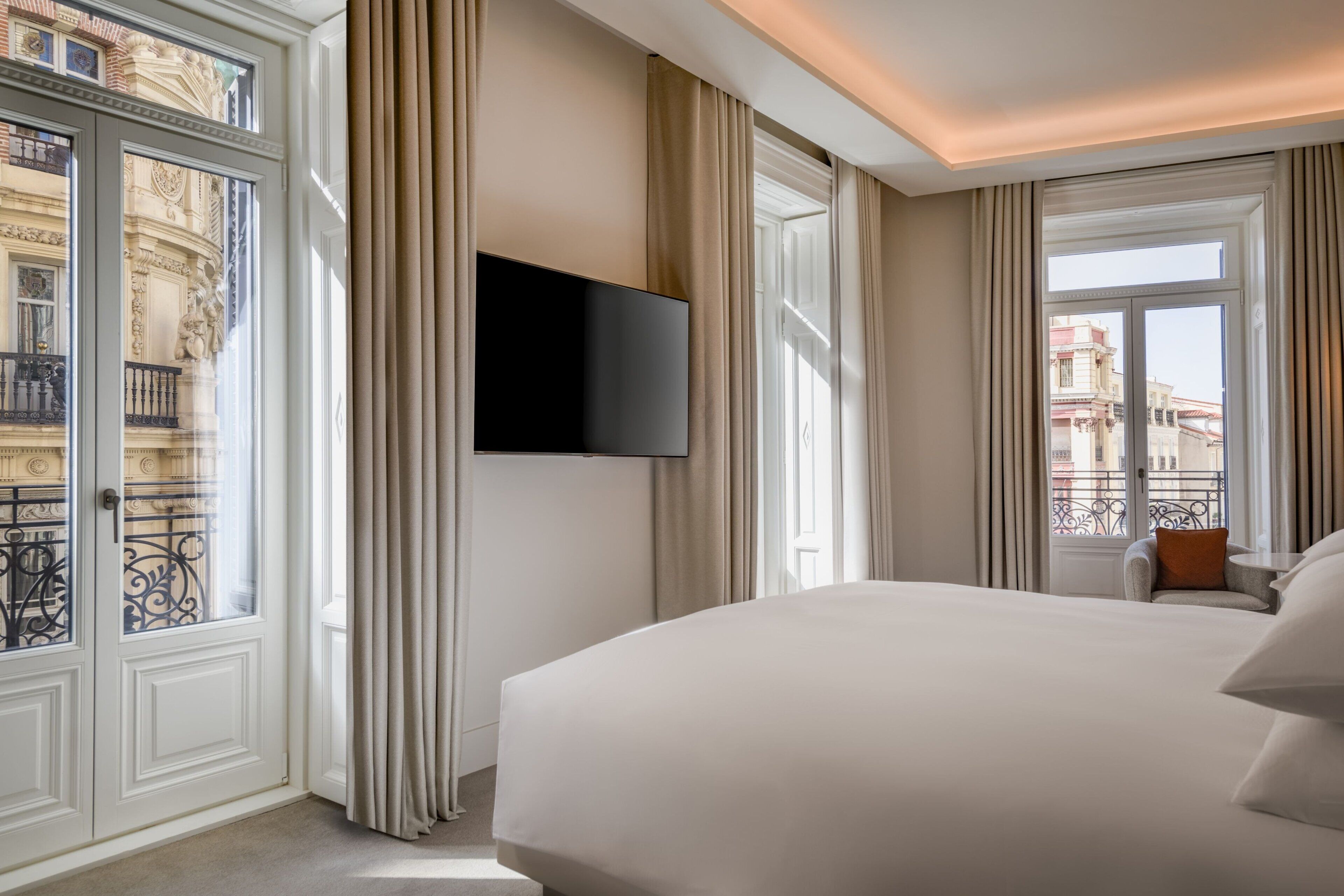 Habitación de hotel con una cama grande, un televisor montado en la pared y vistas de la ciudad a través de ventanas amplias.