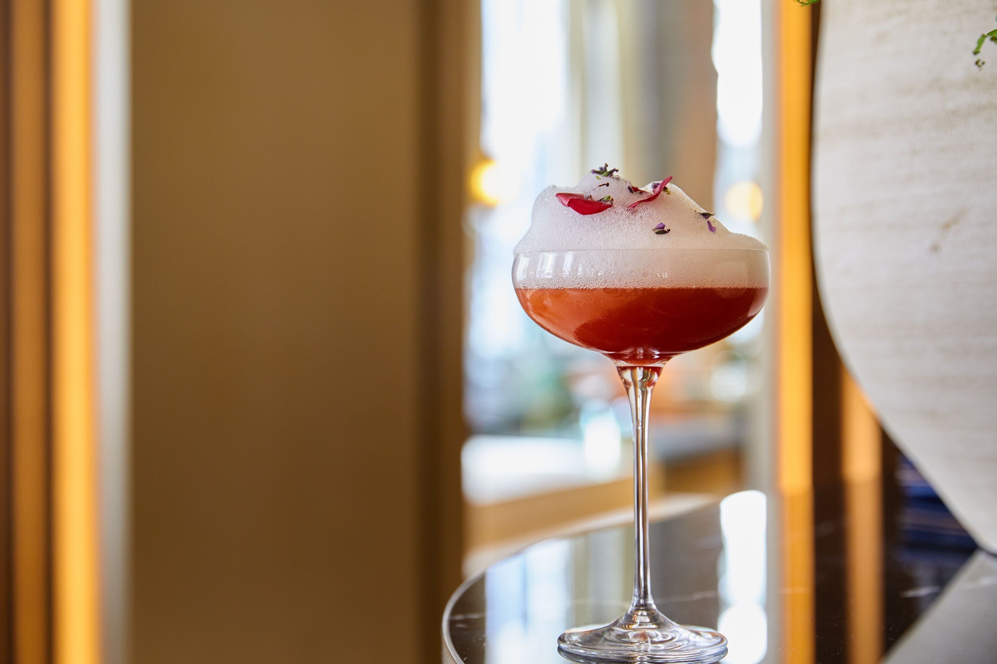 A cocktail in a glass with a frothy top, garnished with herbs, on a reflective surface.