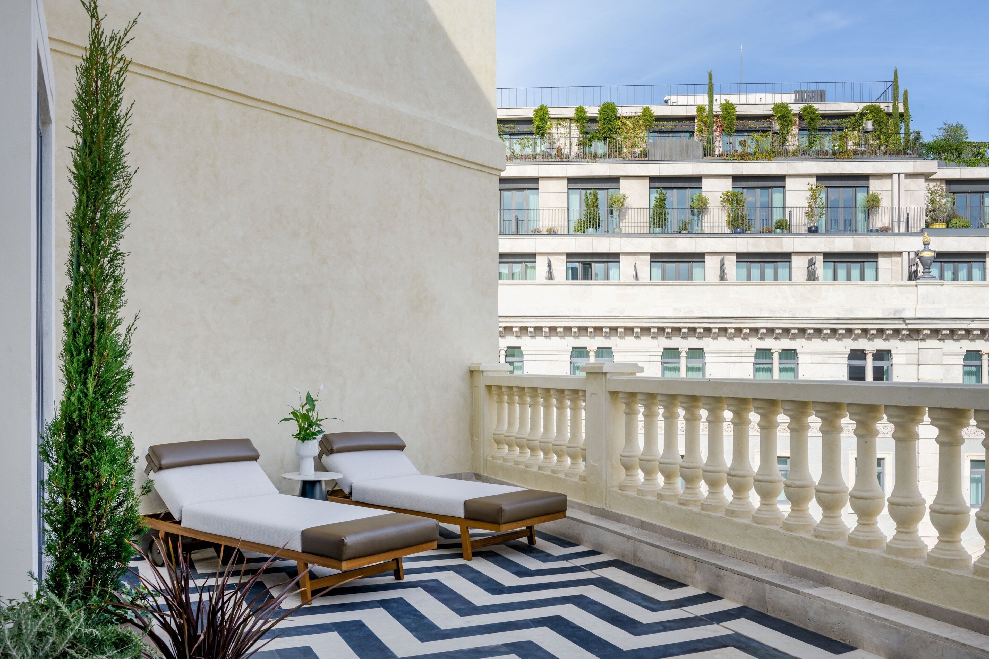 A balcony with two lounge chairs and a small table, overlooking a building with multiple floors and greenery.