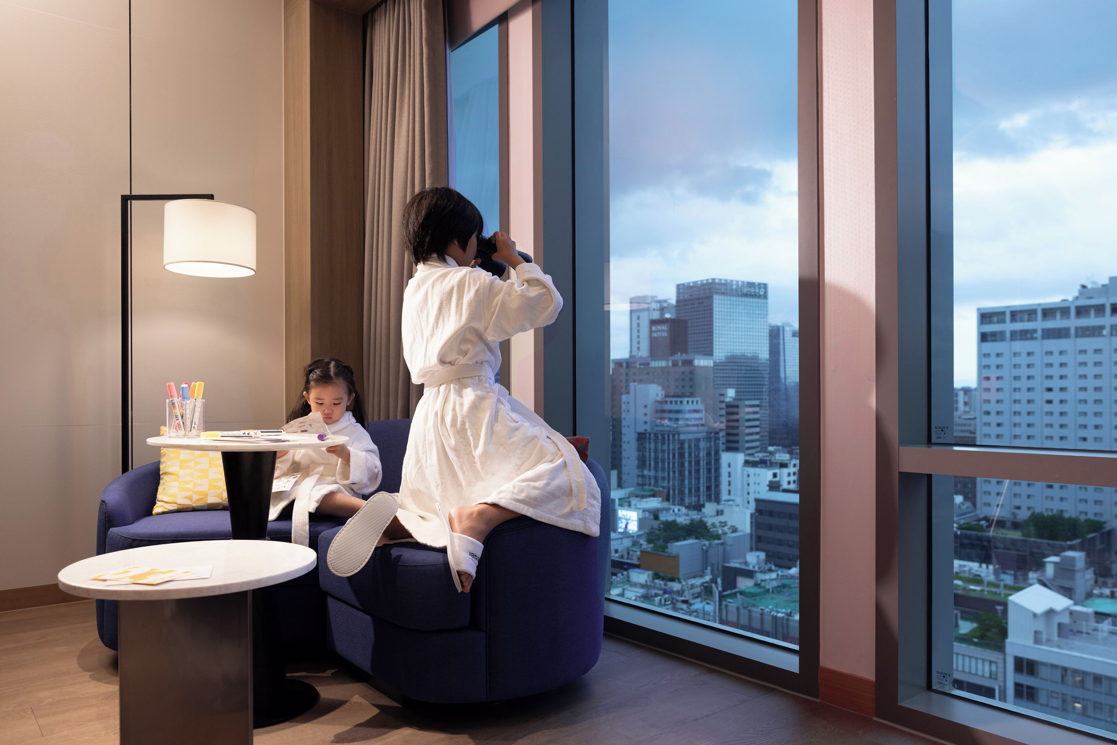 A modern hotel room with a large window offering a city view, a round table with a lamp, and a sofa.