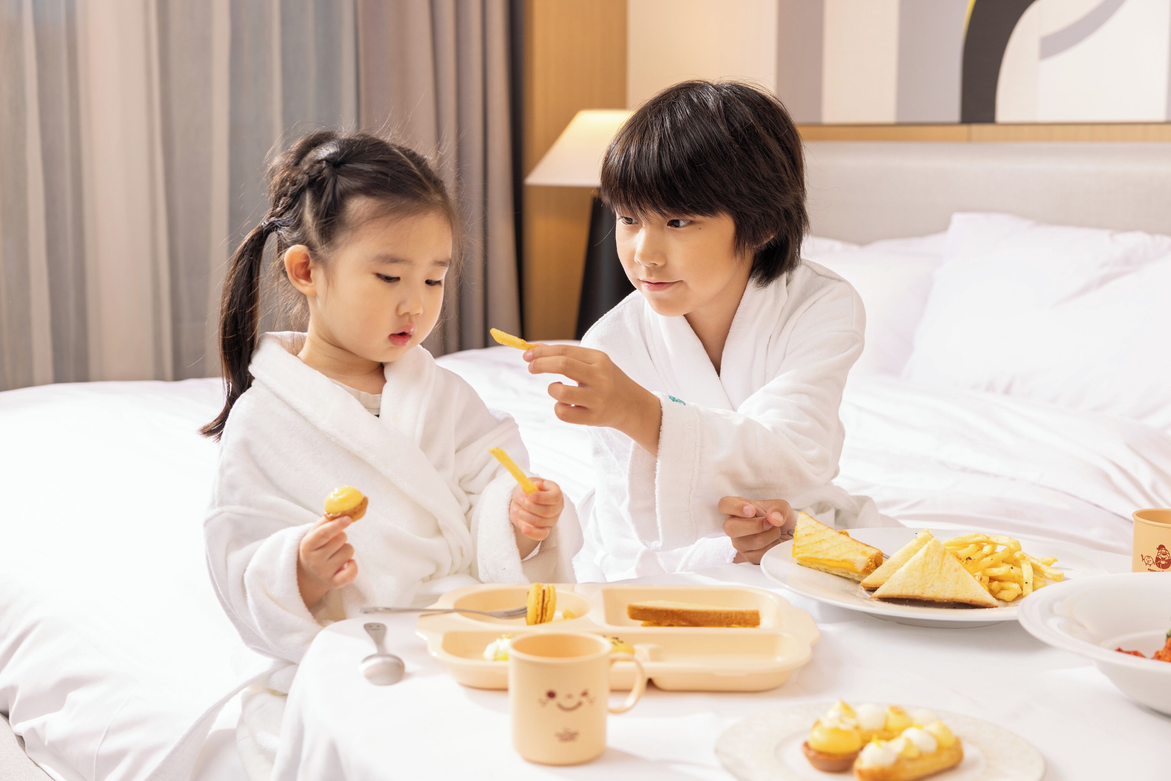 Two individuals in white robes sitting on a bed with a tray of food, including toast and macarons, and a mug with a smiley face.