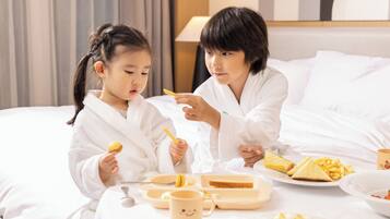 Two individuals in white robes sitting on a bed with a tray of food, including toast and macarons, and a mug with a smiley face.