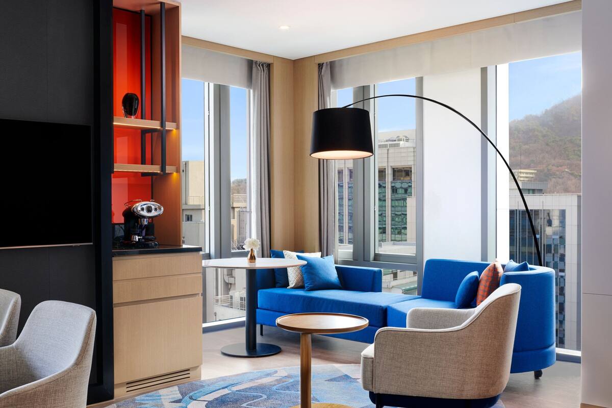 A modern hotel room with a blue sofa, a round coffee table, and a large window offering a city view.