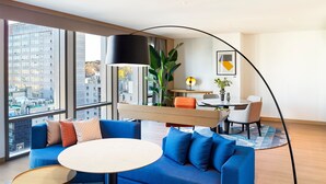 Una habitación de hotel moderna con un sofá azul, una mesa redonda y un área de comedor con una l ámpara y una planta.