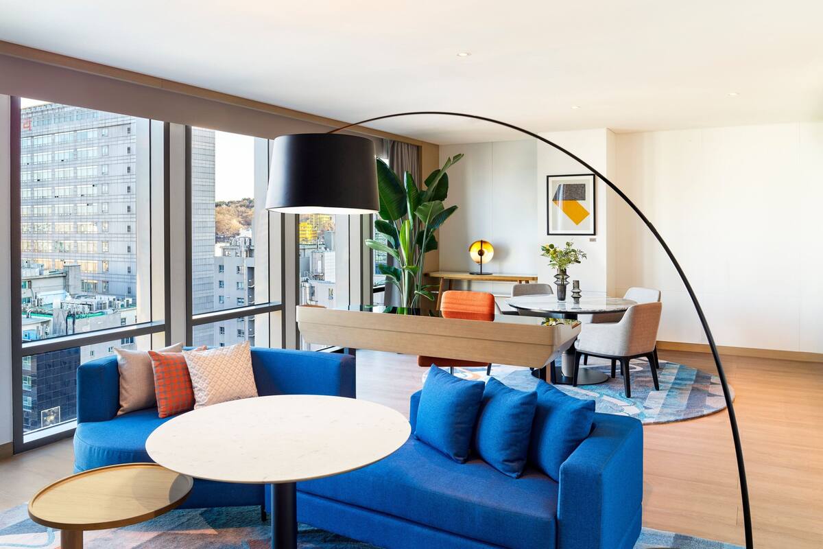 A modern hotel room with a blue sofa, a round table, and a dining area with a lamp and a plant.