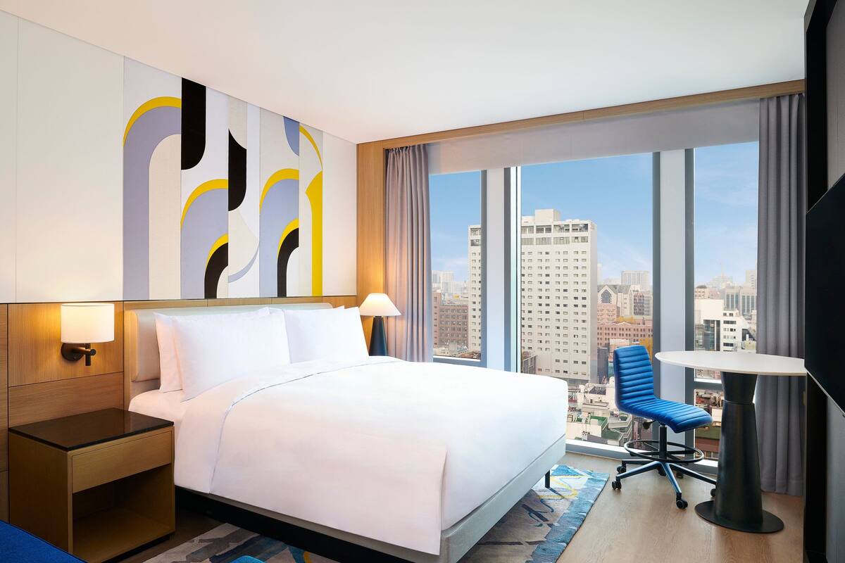 A hotel room with a large bed, a desk with a chair, and a view of the cityscape through large windows.