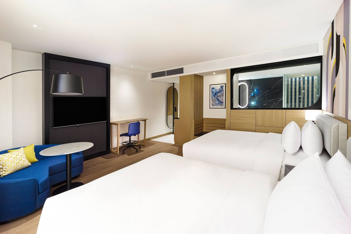 A modern hotel room with a curved blue sofa, a small round table, a desk with a chair, and a large bed with white bedding.