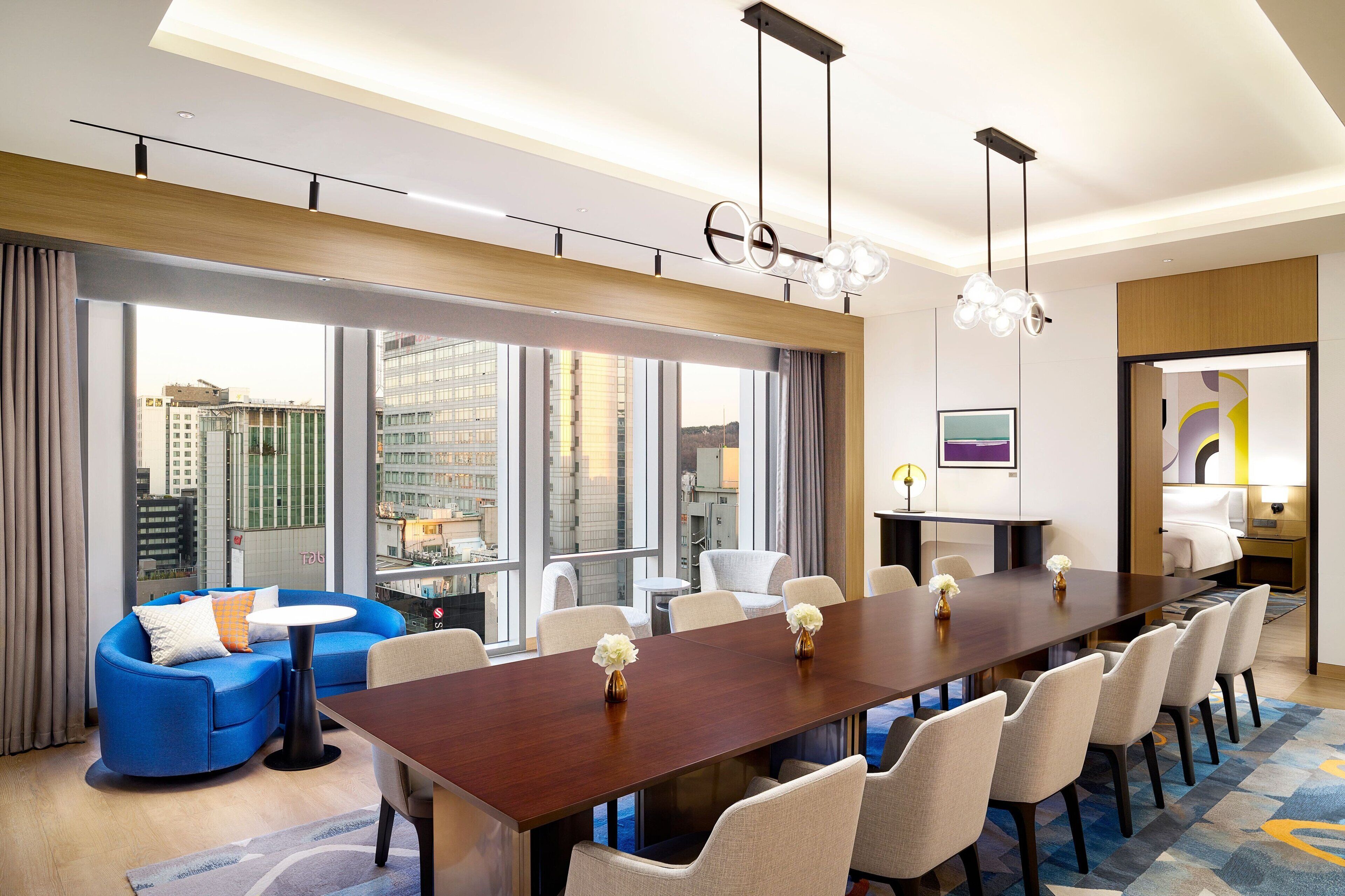 Un salón de conferencias moderno en un hotel, con una larga mesa, varias sillas, un sofá y grandes ventanales con vistas a la ciudad.