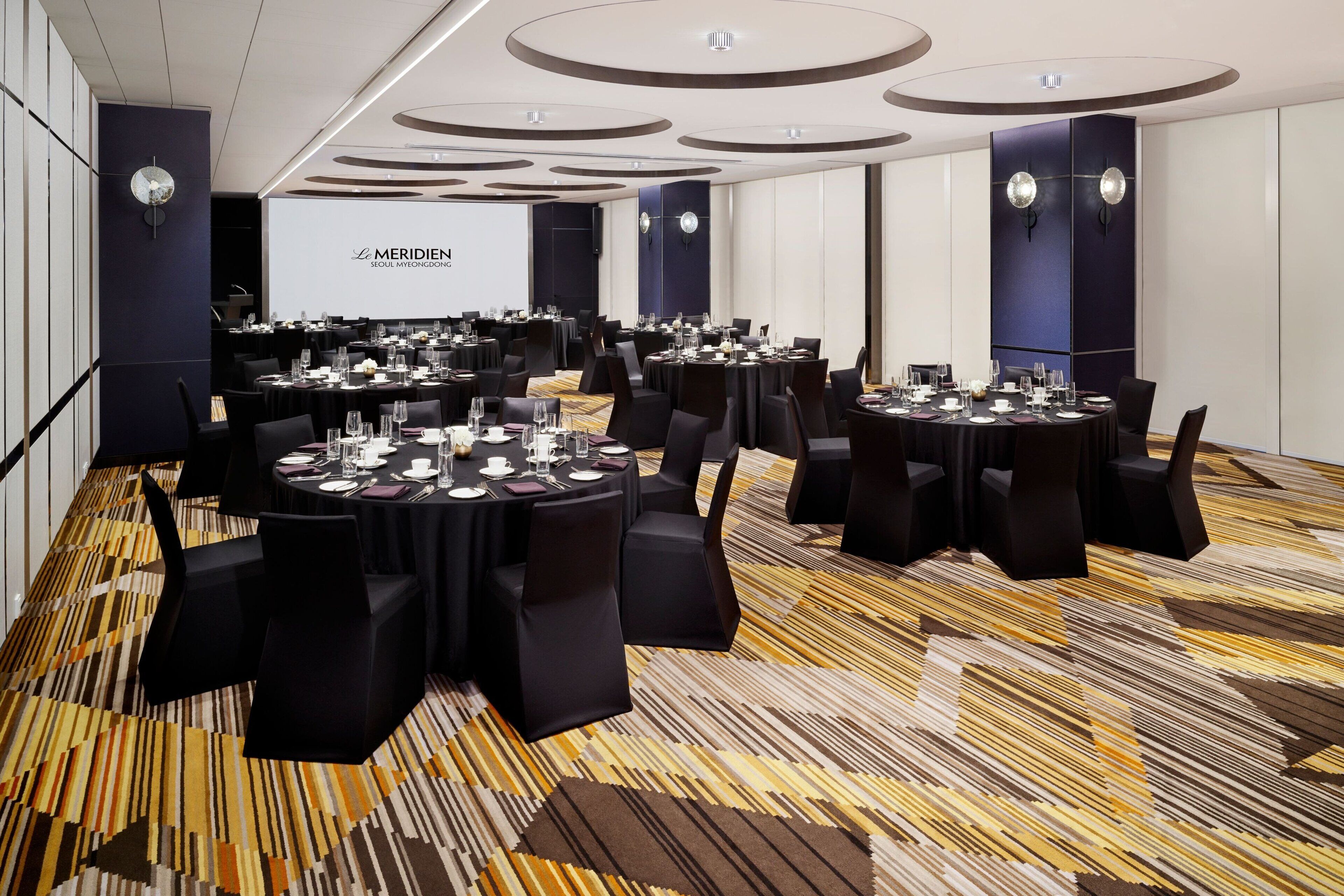 Un salón de conferencias con varias mesas dispuestas para una reunión, equipado con sillas, mesas y pantallas de presentación.
