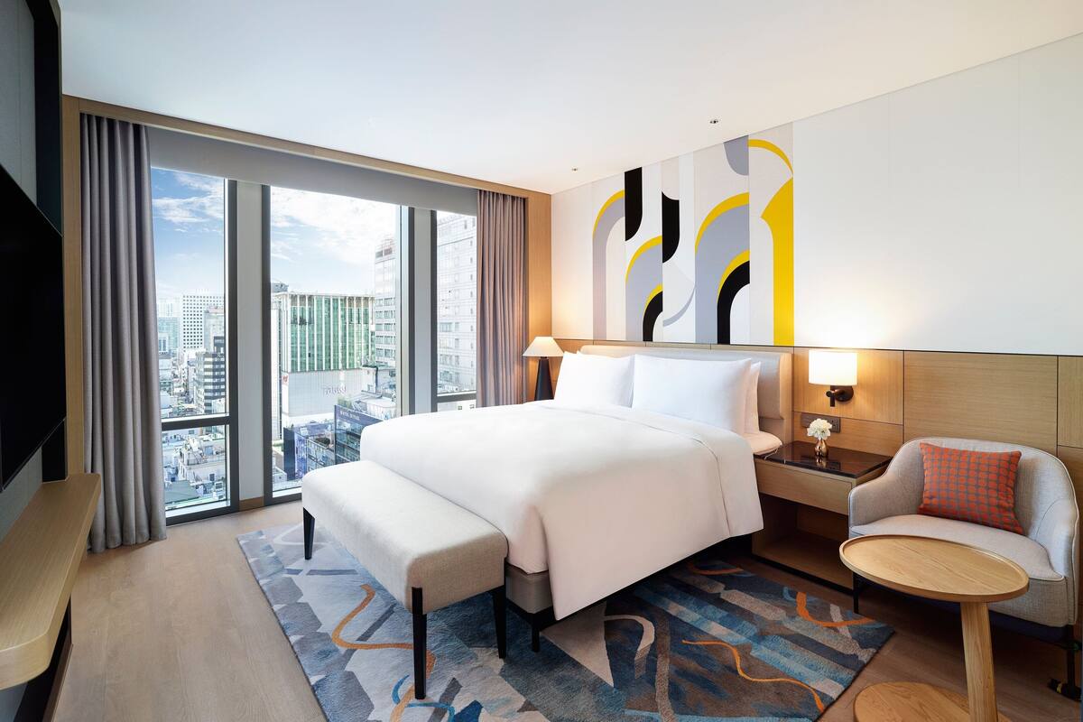 A modern hotel room with a large bed, a small round table, a bench, and a city view through large windows.