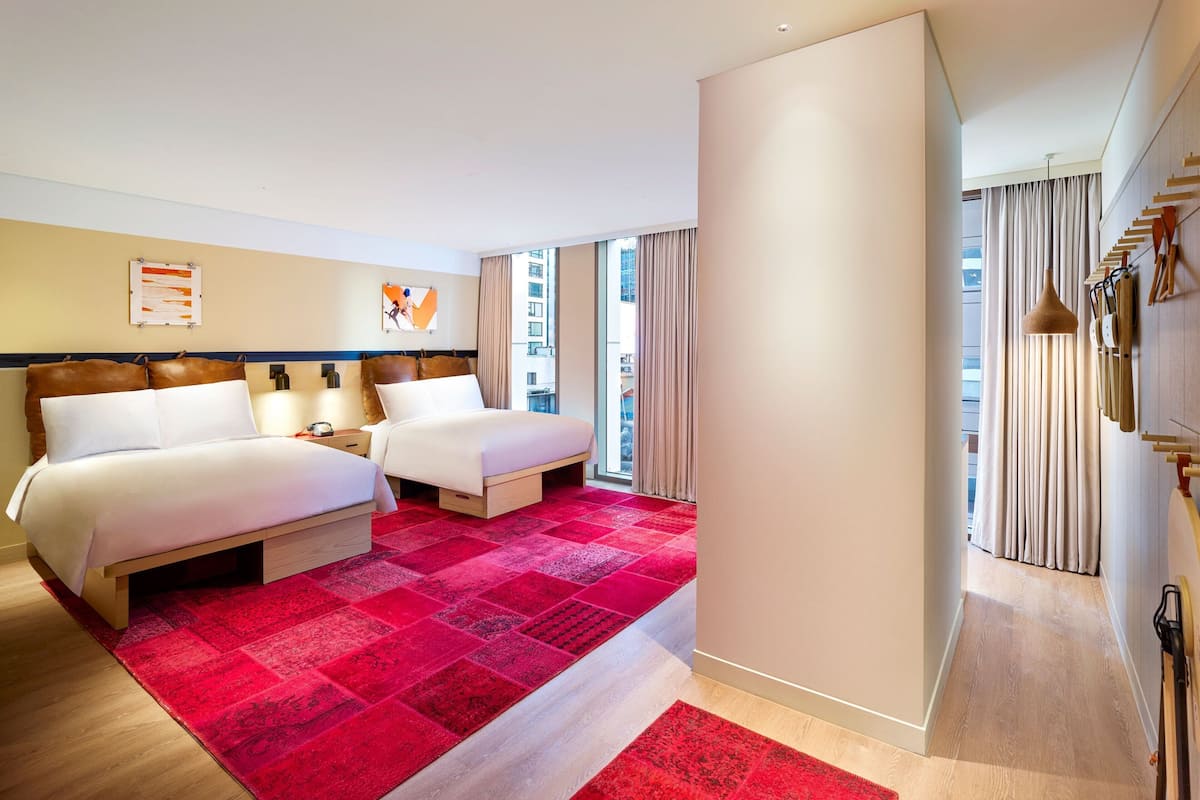 Habitación de hotel con dos camas, una alfombra roja y vistas de la ciudad a través de amplios ventanales.