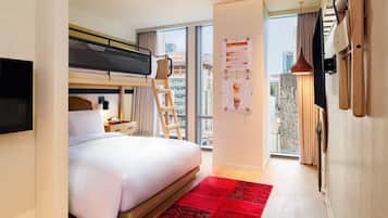 Una habitación de hotel moderna con una litera, un escritorio con lámpara, un tapete rojo y un ventanal con vistas a la ciudad.
