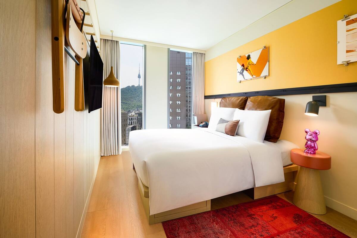 Habitación de hotel con una cama grande, una alfombra roja, una mesita de noche con una lámpara y vistas a un paisaje urbano.
