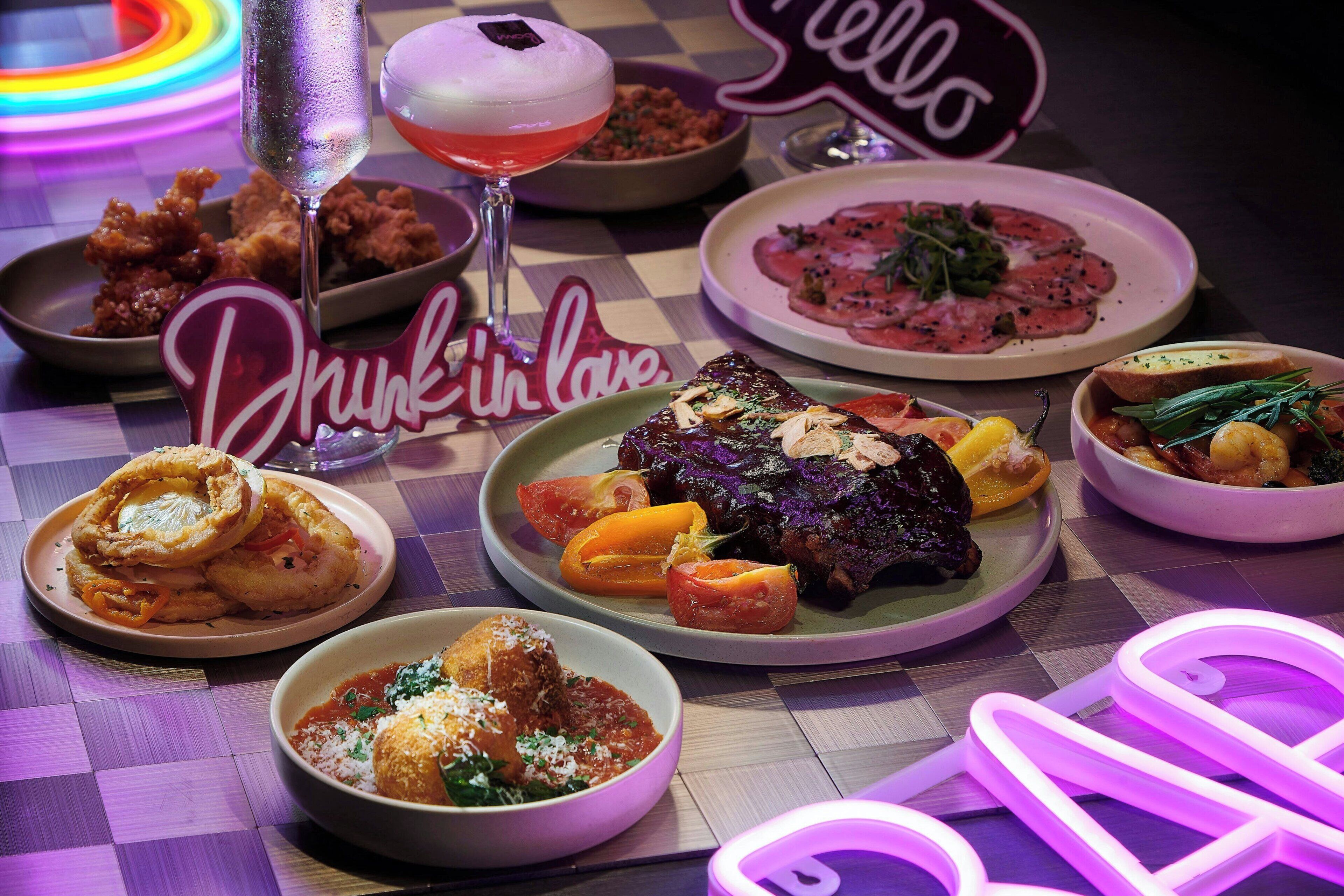 Variedad de platos sobre una mesa, incluyendo un bistec, verduras y alimentos fritos, con un letrero de neón que dice 'Hello' al fondo.