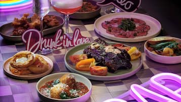 Variedad de platos sobre una mesa, incluyendo un bistec, verduras y alimentos fritos, con un letrero de neón que dice 'Hello' al fondo.