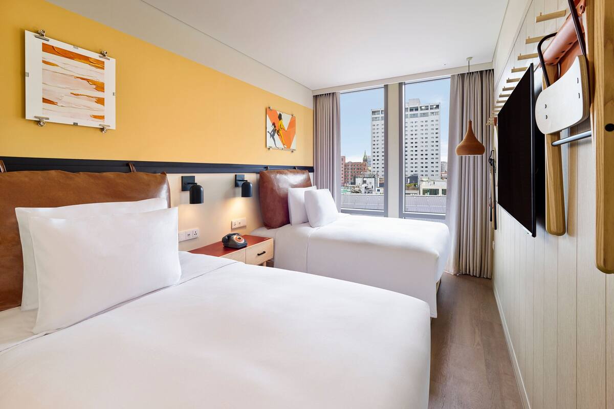 Una habitación de hotel con dos camas, un ventanal con vistas a la ciudad, un televisor de pantalla plana y un cabecero de madera.