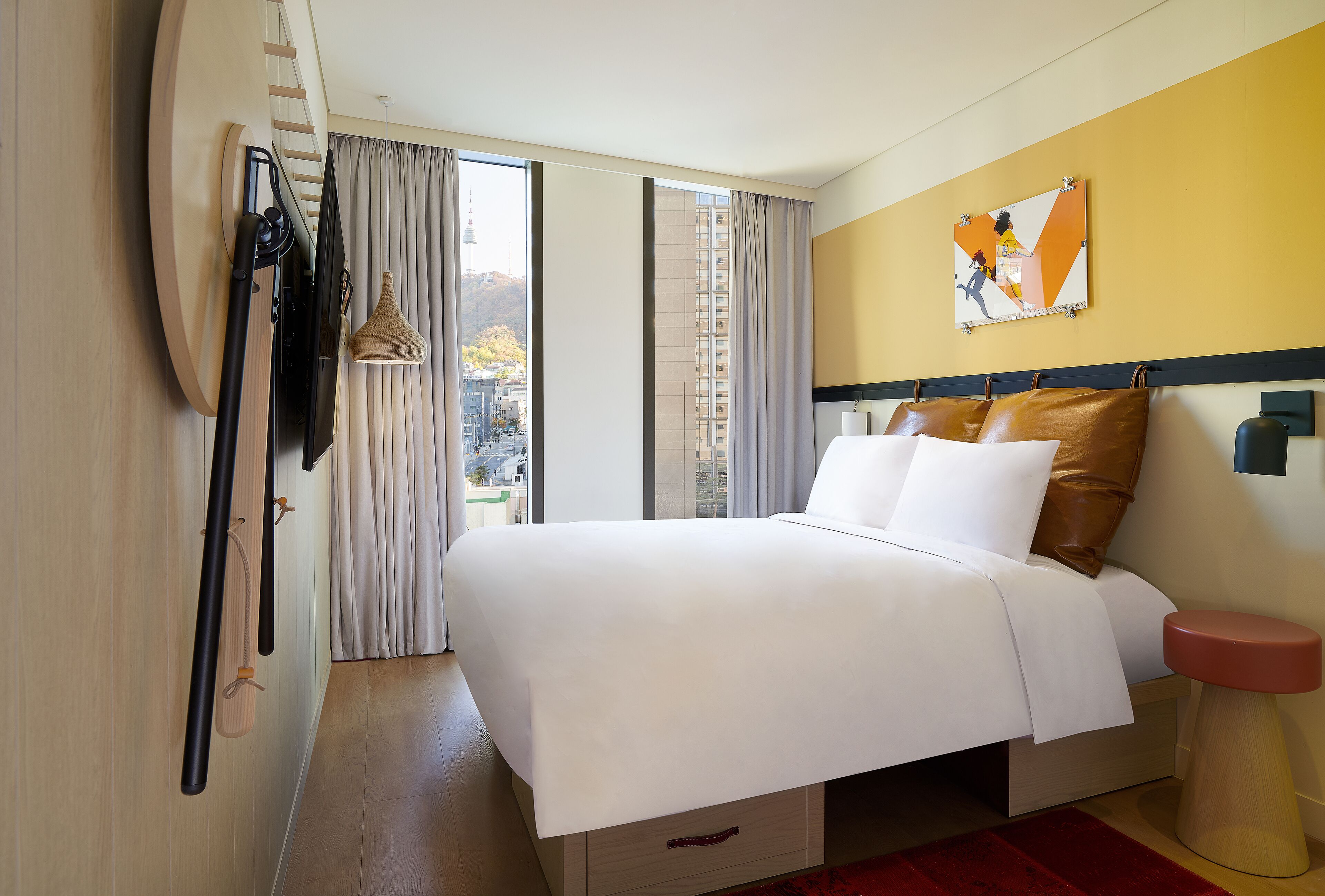 A hotel room with a bed, a window with curtains, a view of a cityscape, and a wall-mounted artwork.
