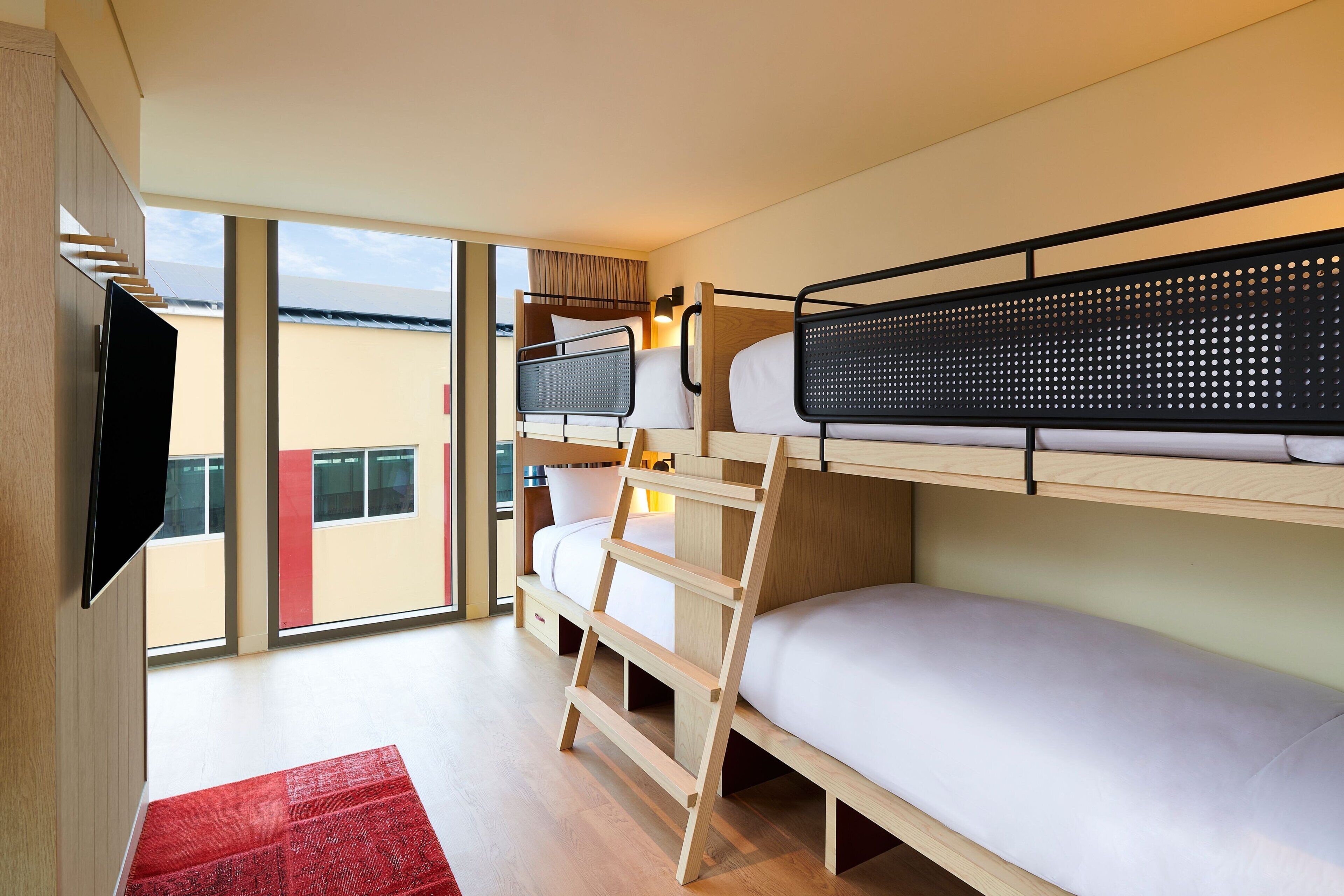 A modern hotel room with a bunk bed, a TV mounted on the wall, and a large window offering a view of the outside.