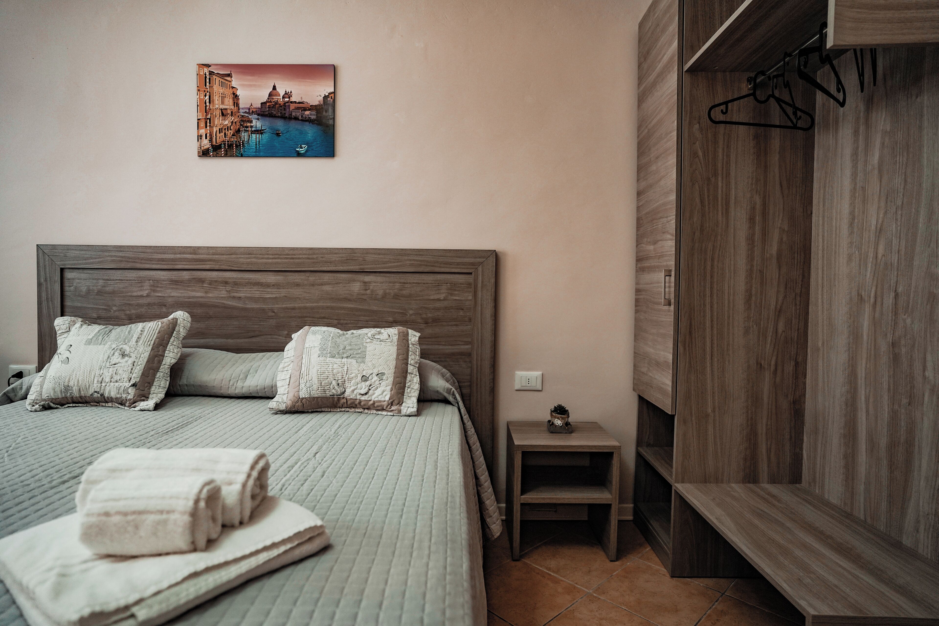 A neatly made bed with pillows, a small bedside table with a plant, and a wooden wardrobe with a hanger.