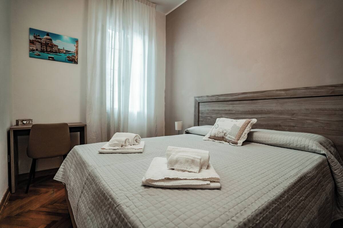 A hotel room with a bed, a desk, and a chair. There is a painting on the wall and a window with curtains.