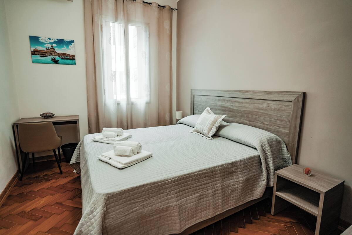 A neatly made bed with a wooden headboard, a small bedside table, and a chair in a room with wooden flooring and a window with curtains.