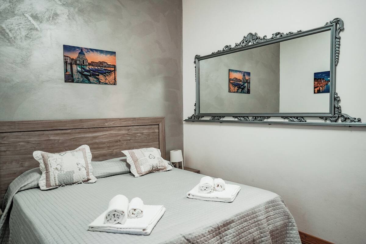 A neatly made bed with pillows, a folded towel, and a mirror with a picture of a cityscape.