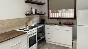 A compact kitchen with a stove, oven, white cabinets, a small sink, and a window with blinds. - Town Beach Short Stay (Mandurah)