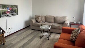 A living room with a beige sofa, a small round table, a television mounted on the wall, and a wooden floor. - Town Beach Short Stay (Mandurah)