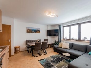 A modern living room with a grey sofa, a dining table with four chairs, a flat-screen TV mounted on the wall, and a window with decorative items.