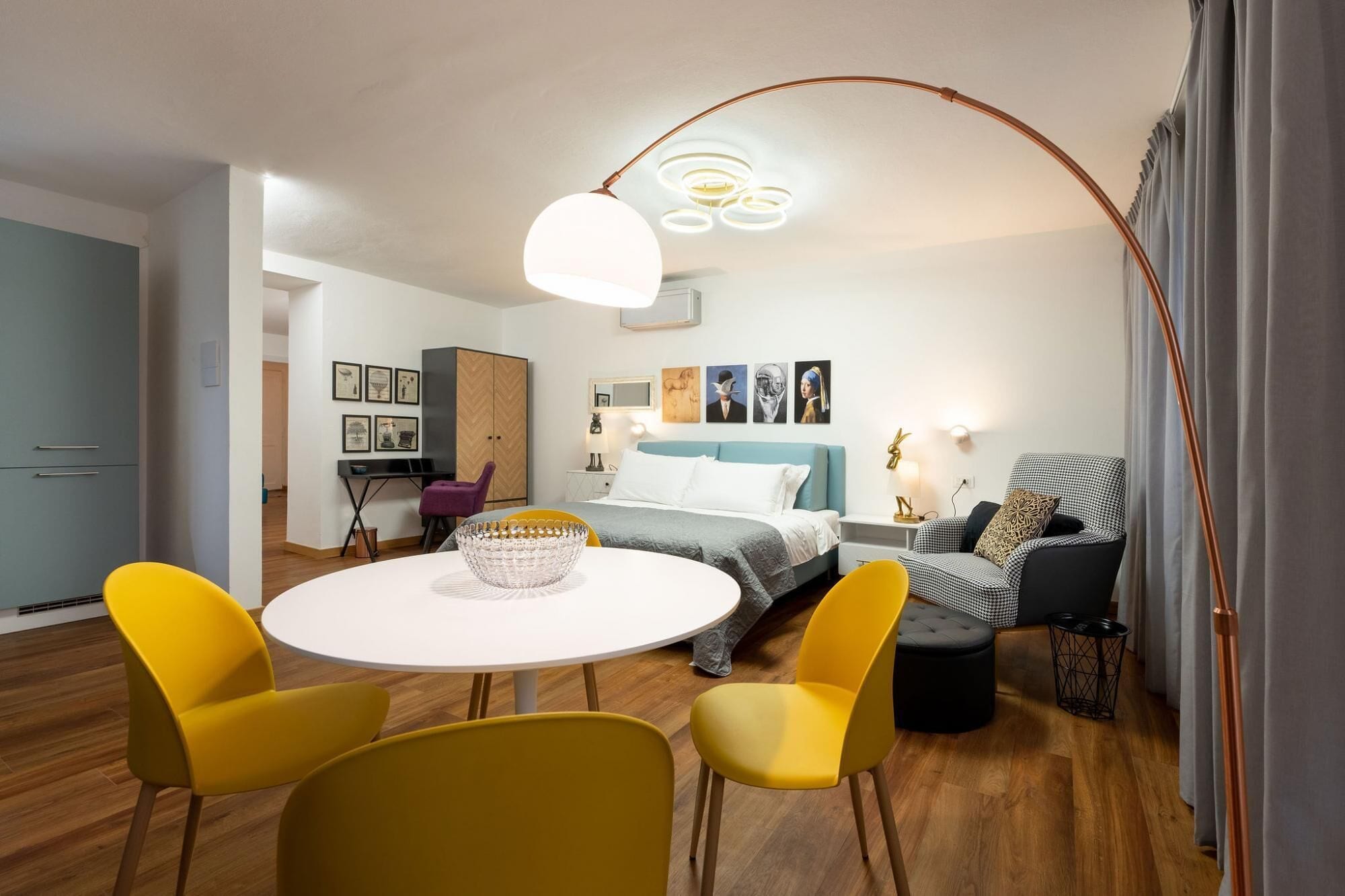 A modern, well-lit room with a round dining table, yellow chairs, a bed with a blue headboard, and a herringbone-patterned cabinet.