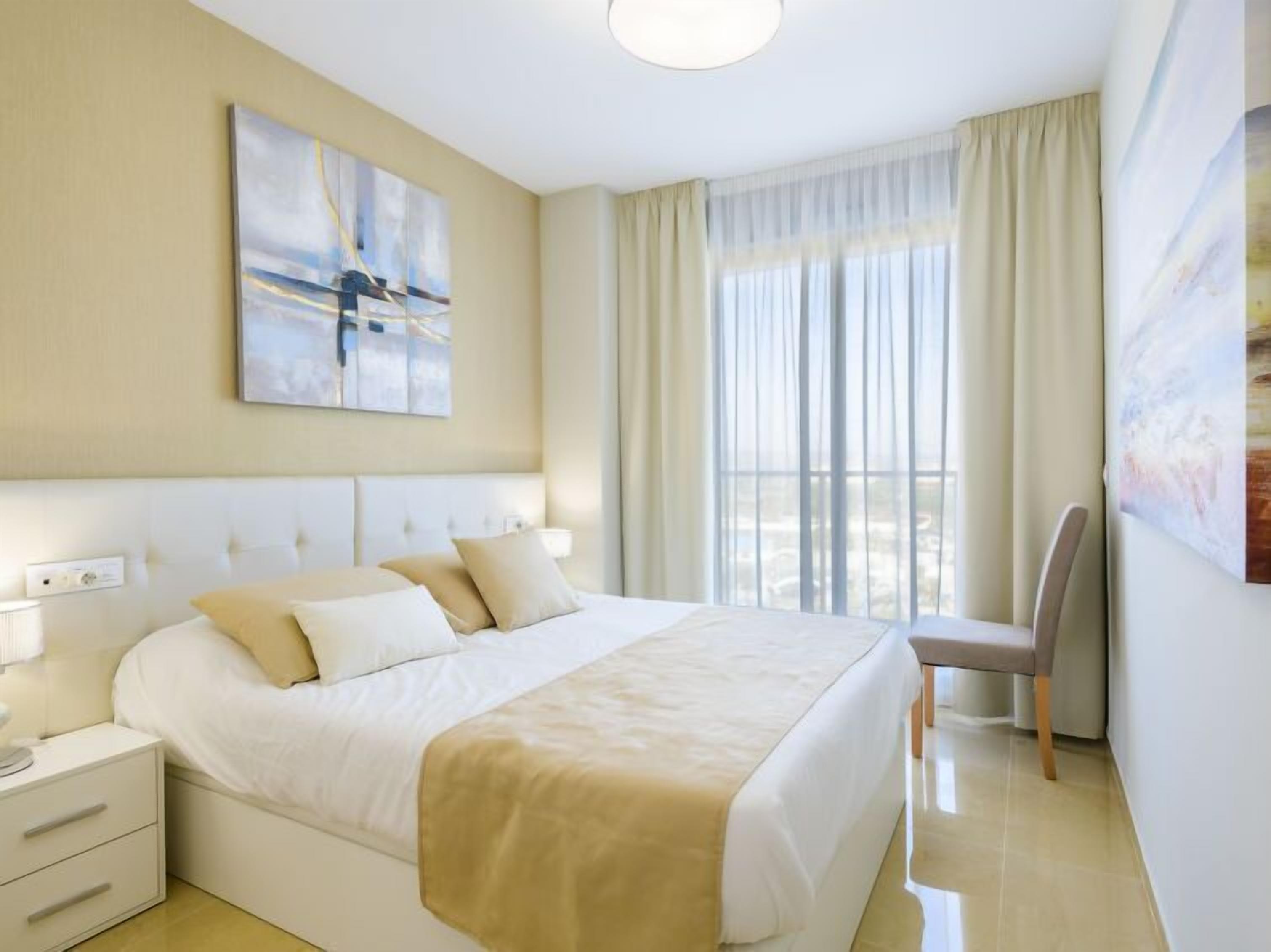 Habitación de hotel con una cama grande, una mesita de noche, una silla y un ventanal con cortinas.