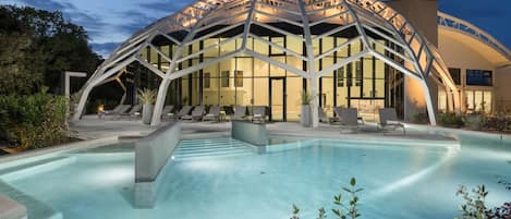 A modern hotel with a large outdoor pool, lounge chairs, and a distinctive arched glass facade.