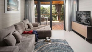 Ein modernes Wohnzimmer mit grauem Sofa, einem Couchtisch und einem Fernseher auf einer Kommode. Ein Balkon mit Holzboden und Vorhängen ist ebenfalls vorhanden.