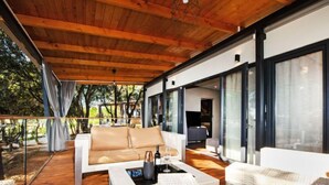 Ein moderner Außenbereich mit Holzterrasse, einem weißen Sofa, Korbmöbeln und einem Glasgeländer mit Blick auf einen Garten.