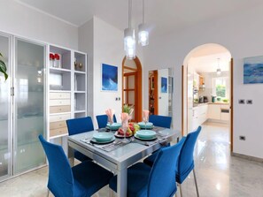 A modern dining area with a glass table, blue chairs, and a kitchen visible through an archway.