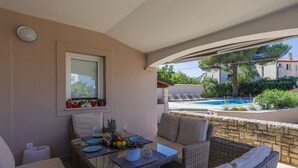 A sunlit patio with wicker furniture, a table set with fruits and glasses, and a view of a swimming pool and greenery.