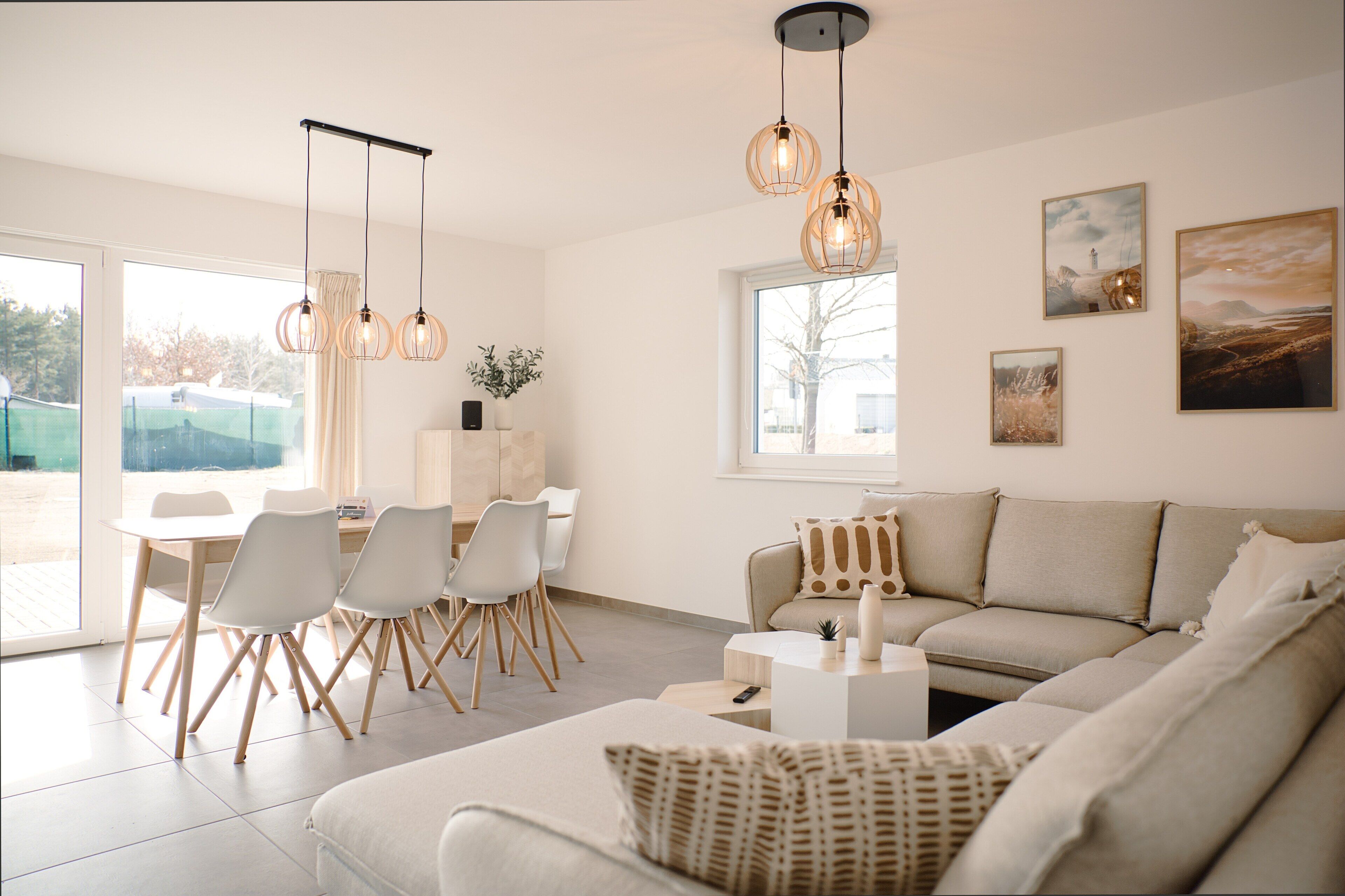 Un salon moderne avec un canapé, une table à manger et des chaises. On y trouve des luminaires suspendus, des œuvres d’art accrochées aux murs et une grande fenêtre.