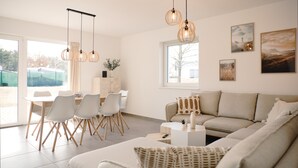 A modern living room with a sofa, dining table, and chairs. There are pendant lights, artwork on the walls, and a large window. - Modern Swedish house Jonte #3 on Lake Bärwald - SKAN-PARK (Boxberg, OT Klitten)